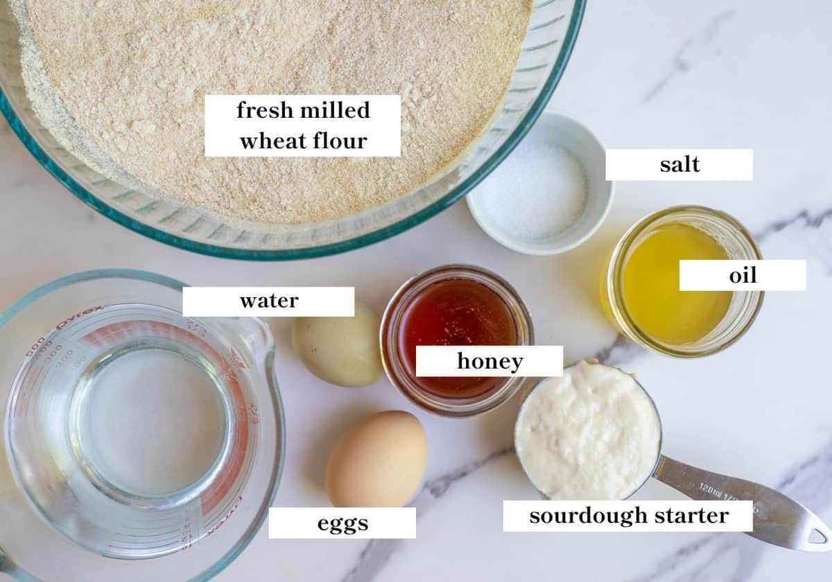 Freshly milled wheat flour, water,  honey, oil, eggs, and more on a countertop.