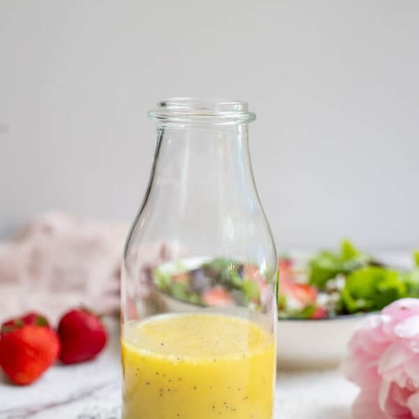 Homemade poppy seed salad dressing in a glass bottle with a salad and strawberries in the background.