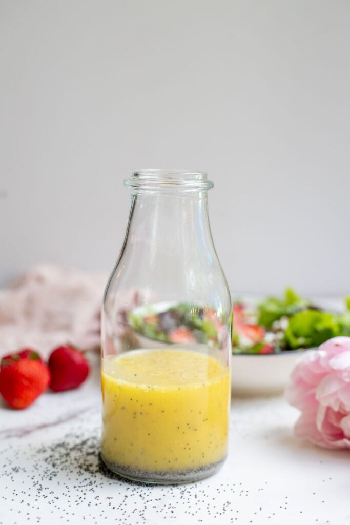 Homemade poppy seed salad dressing in a glass bottle with a salad and strawberries in the background.