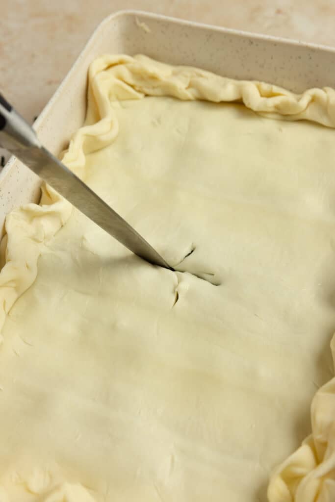 A knife slicing an x into puff pastry dough.