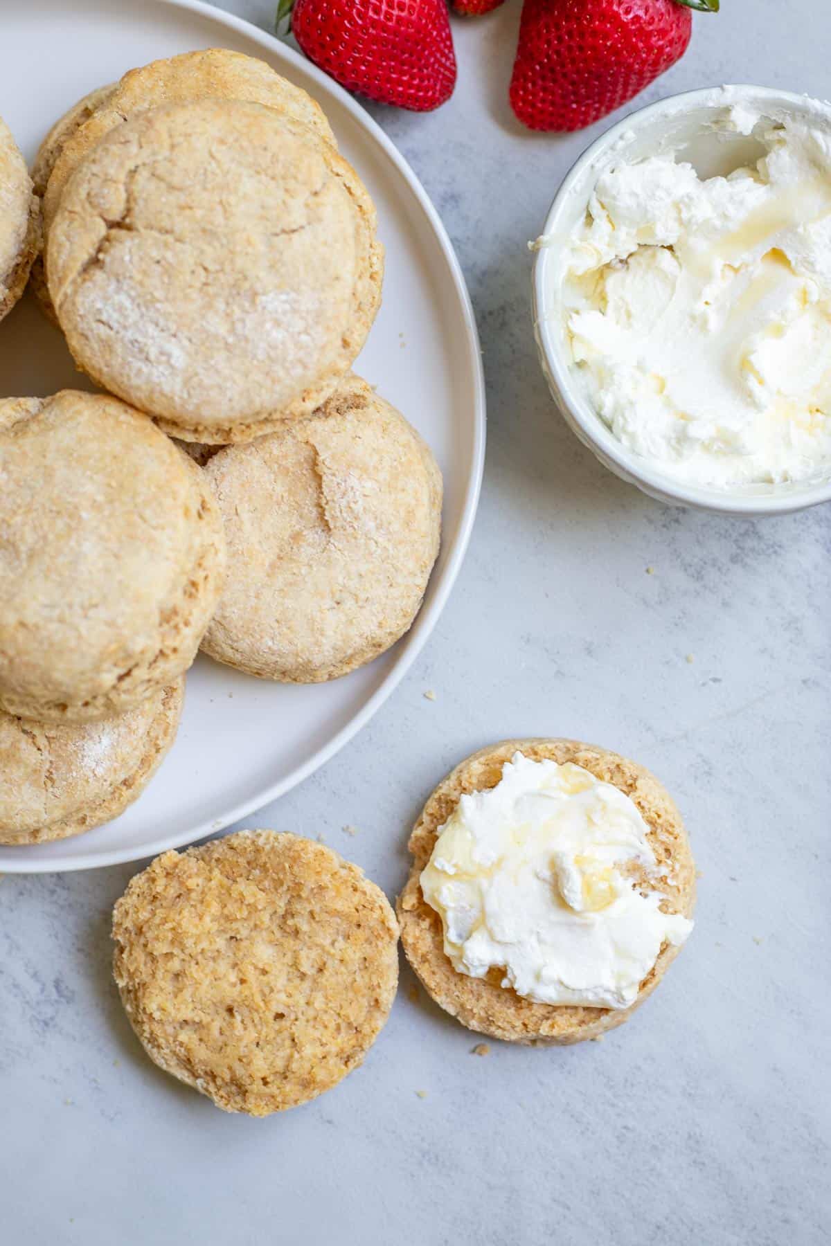 A sliced and buttered whole wheat biscuit beside a plate of biscuits, a bowl of cream cheese, and strawberries.