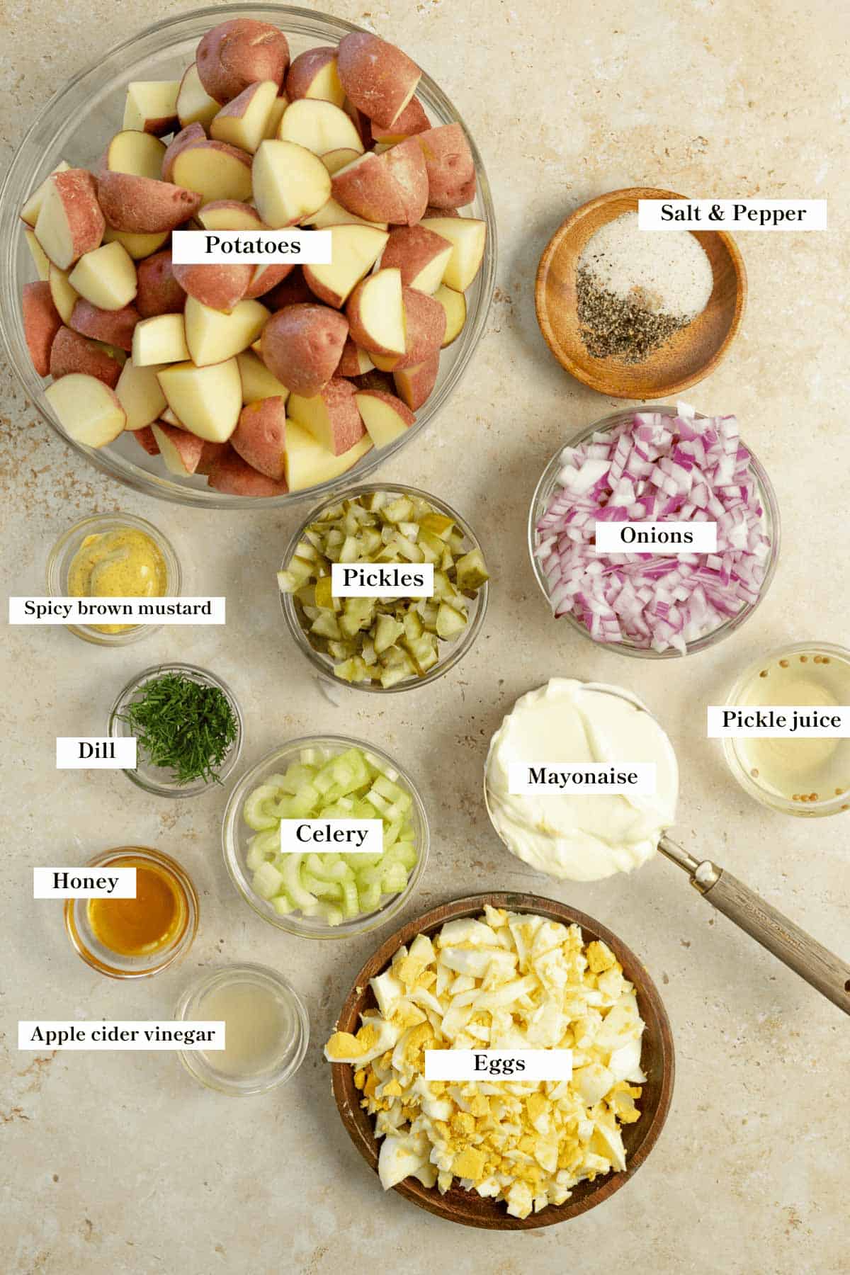 Potatoes, onions, pickles, celery and other ingredients in bowls on a countertop.
