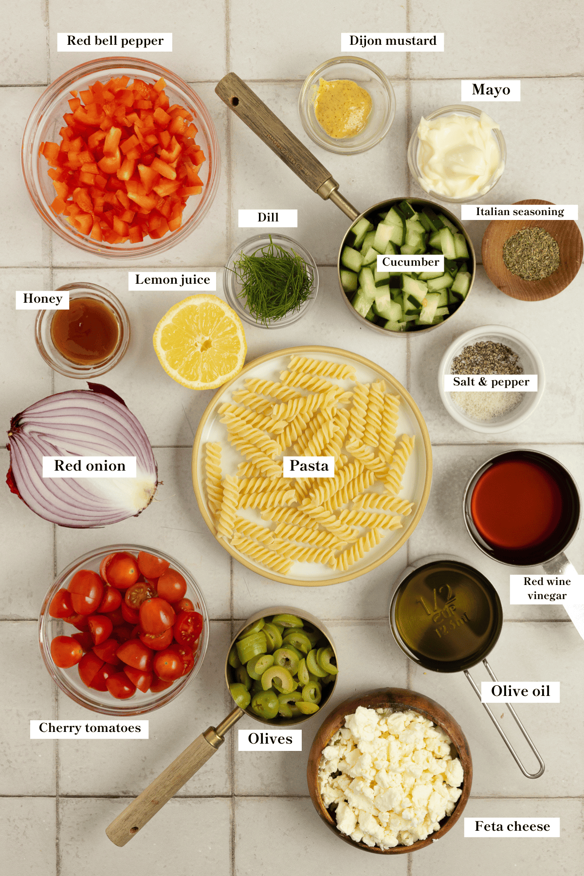 A bowl of pasta, tomatoes, olives, and other ingredients for Greek pasta salad on a countertop.
