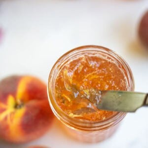 a knife scooping out peach jelly from the glass jar.