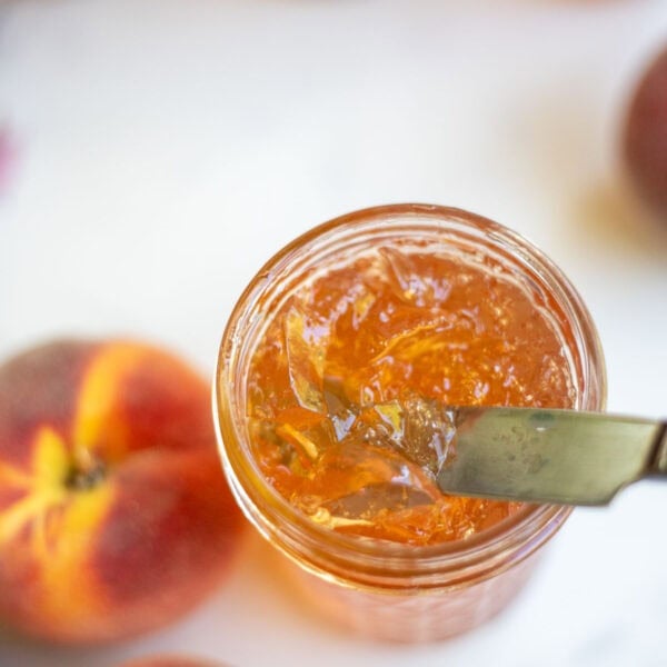 a knife scooping out peach jelly from the glass jar.