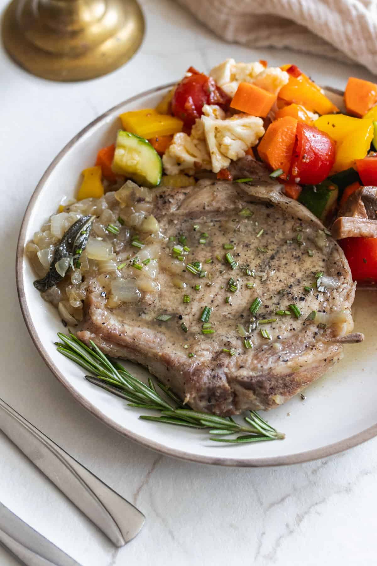 a cooked pork chop in herbs on a white plate with vegetables.