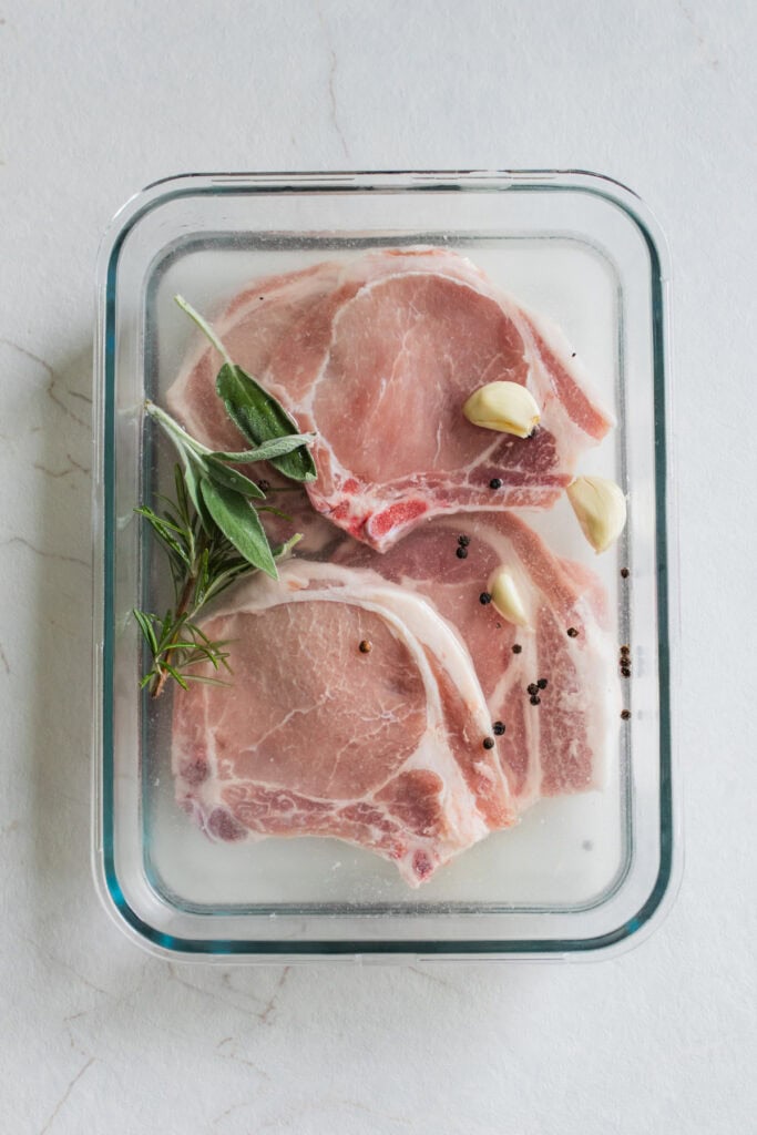 Pork chops brining in a glass container with fresh herbs and garlic.