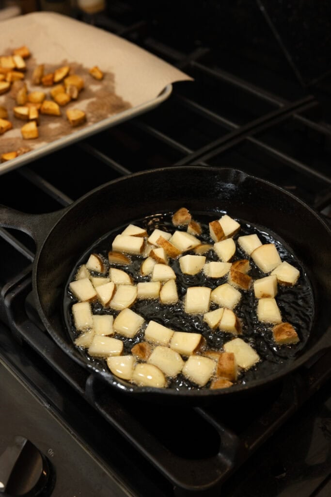 Second batch of potatoes cooking in the skillet, first batch draining in a tray.