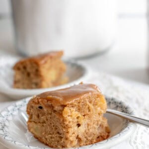 A slice of apple cake topped with caramel topping in a white plate with another slice in the background.
