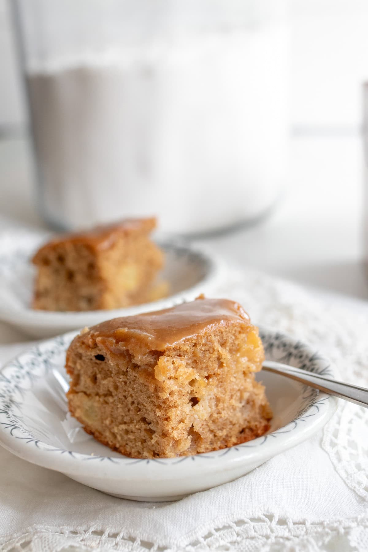 A slice of apple cake topped with caramel topping in a white plate with another slice in the background.