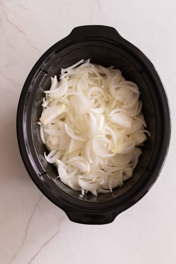 Sliced onions layered in the bottom of a slow cooker.