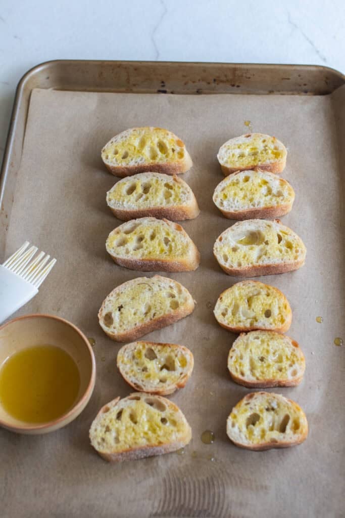 Slices of baguette bread buttered and lining a parchment paper.