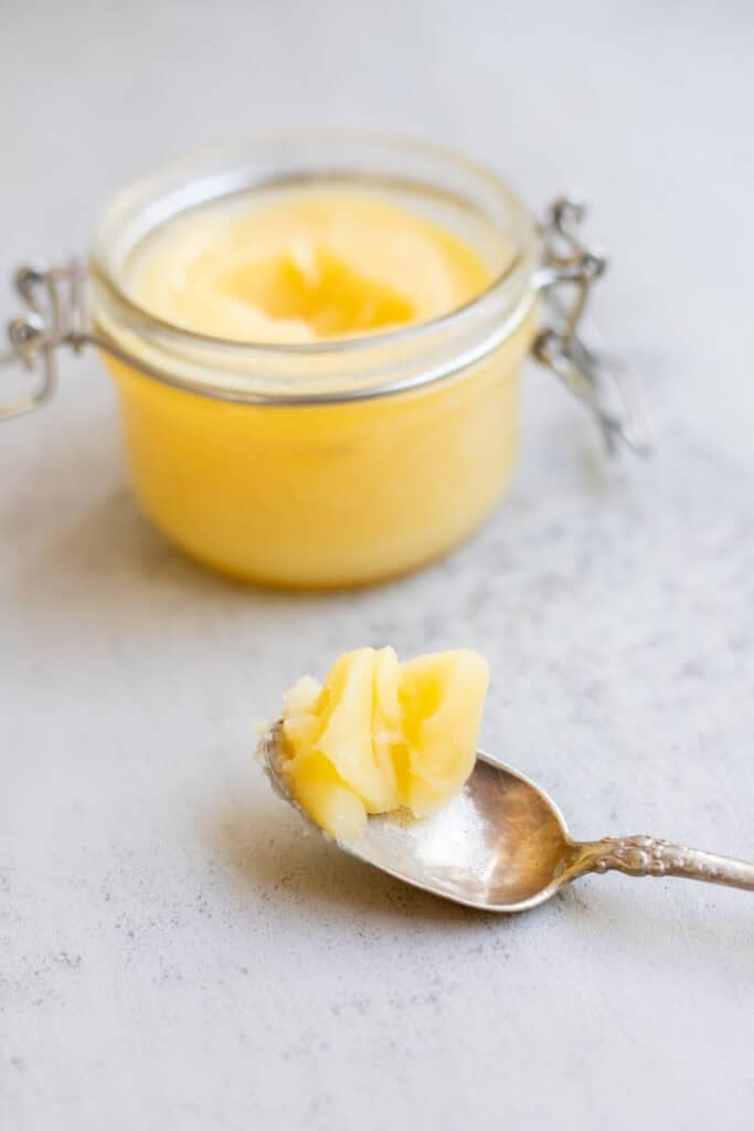 A jar full of cooled ghee with a spoon dipped in ghee laying in front of it. 