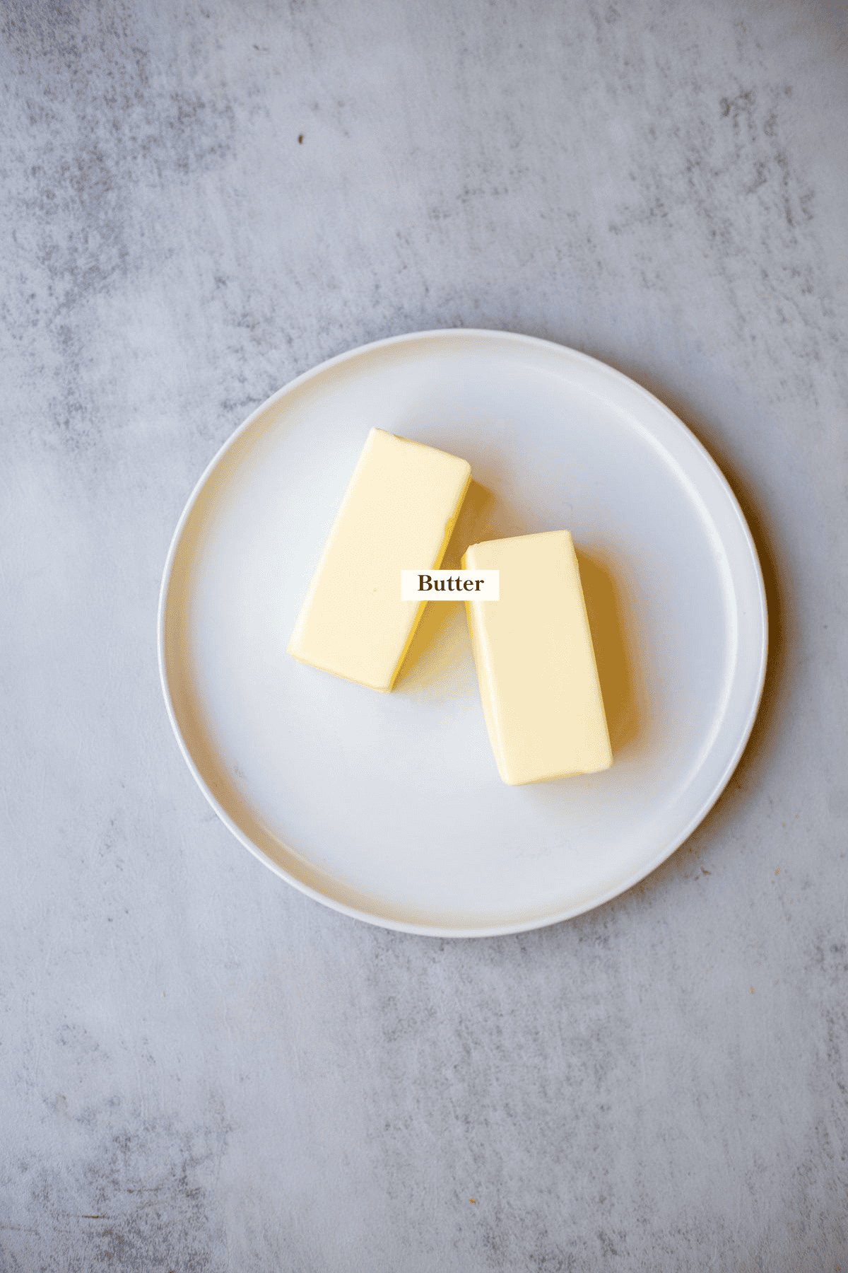 Two sticks of butter on a white plate sitting on a gray countertop.