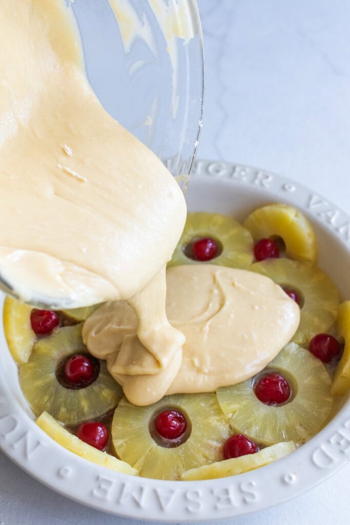 Pouring cake batter over sliced pineapple rings and maraschino cherries.