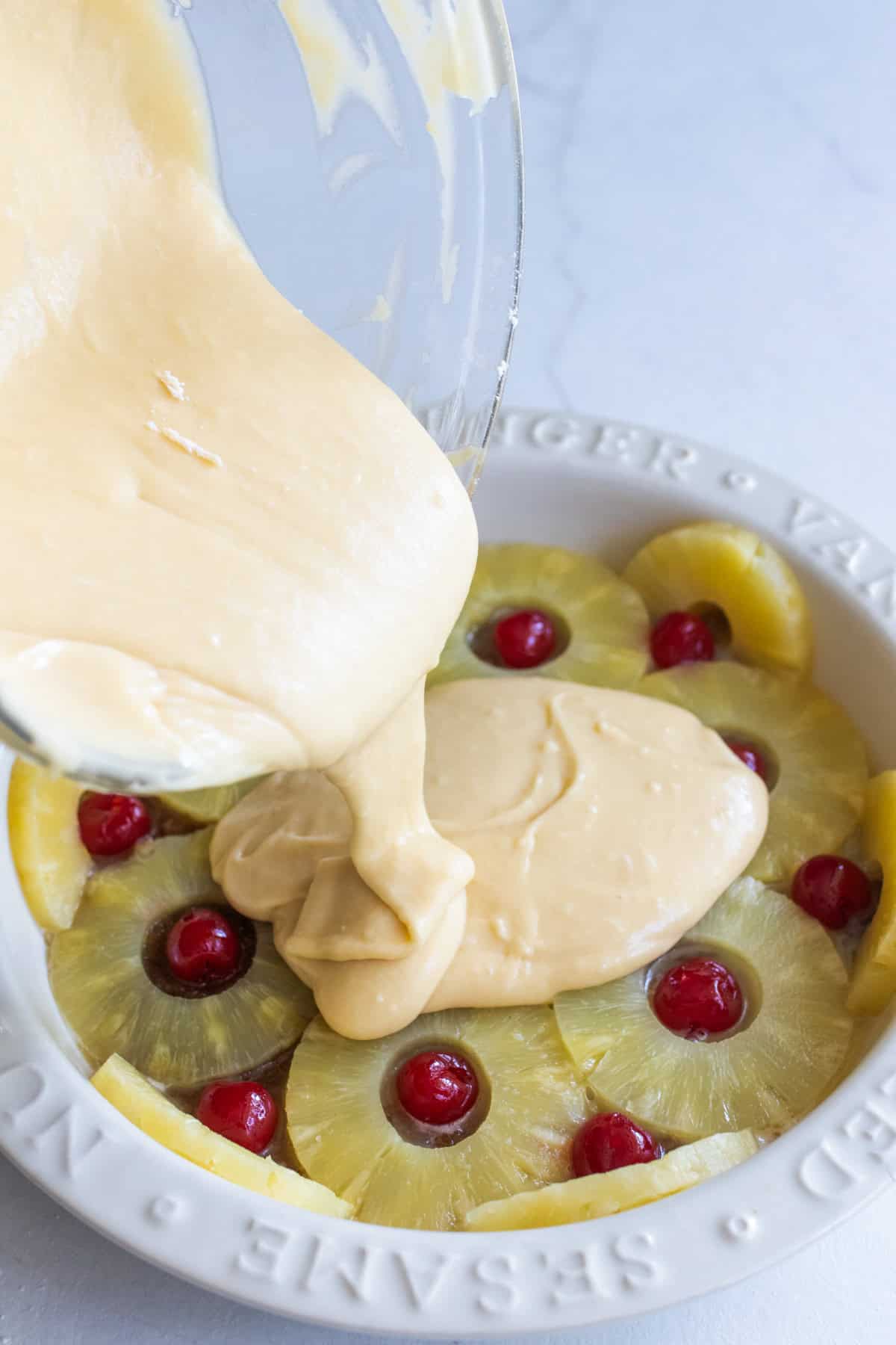 Pouring cake batter over sliced pineapple rings and maraschino cherries.