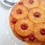 Close up picture of a pineapple upside down cake on a glass cake stand with plates in the background.
