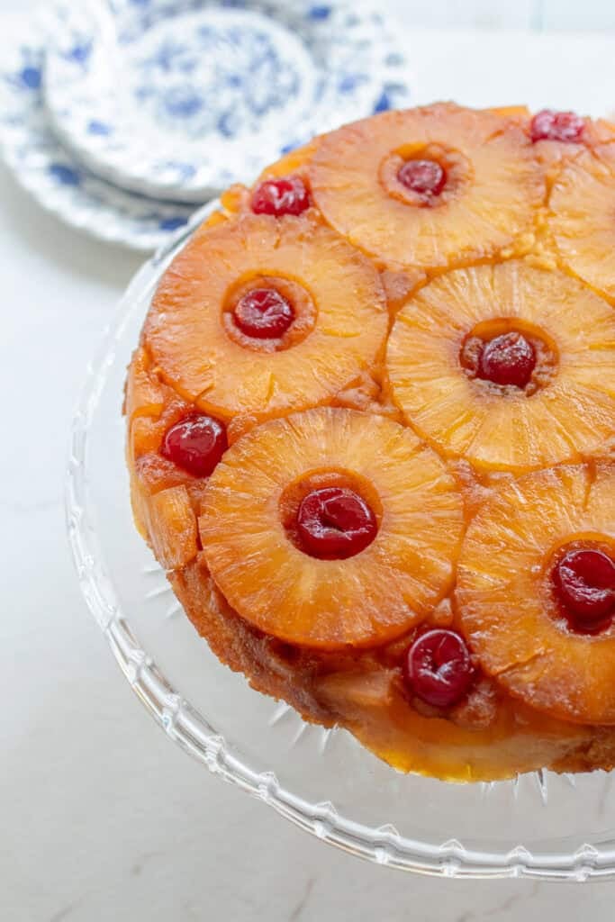 Close up picture of a pineapple upside down cake on a glass cake stand with plates in the background.