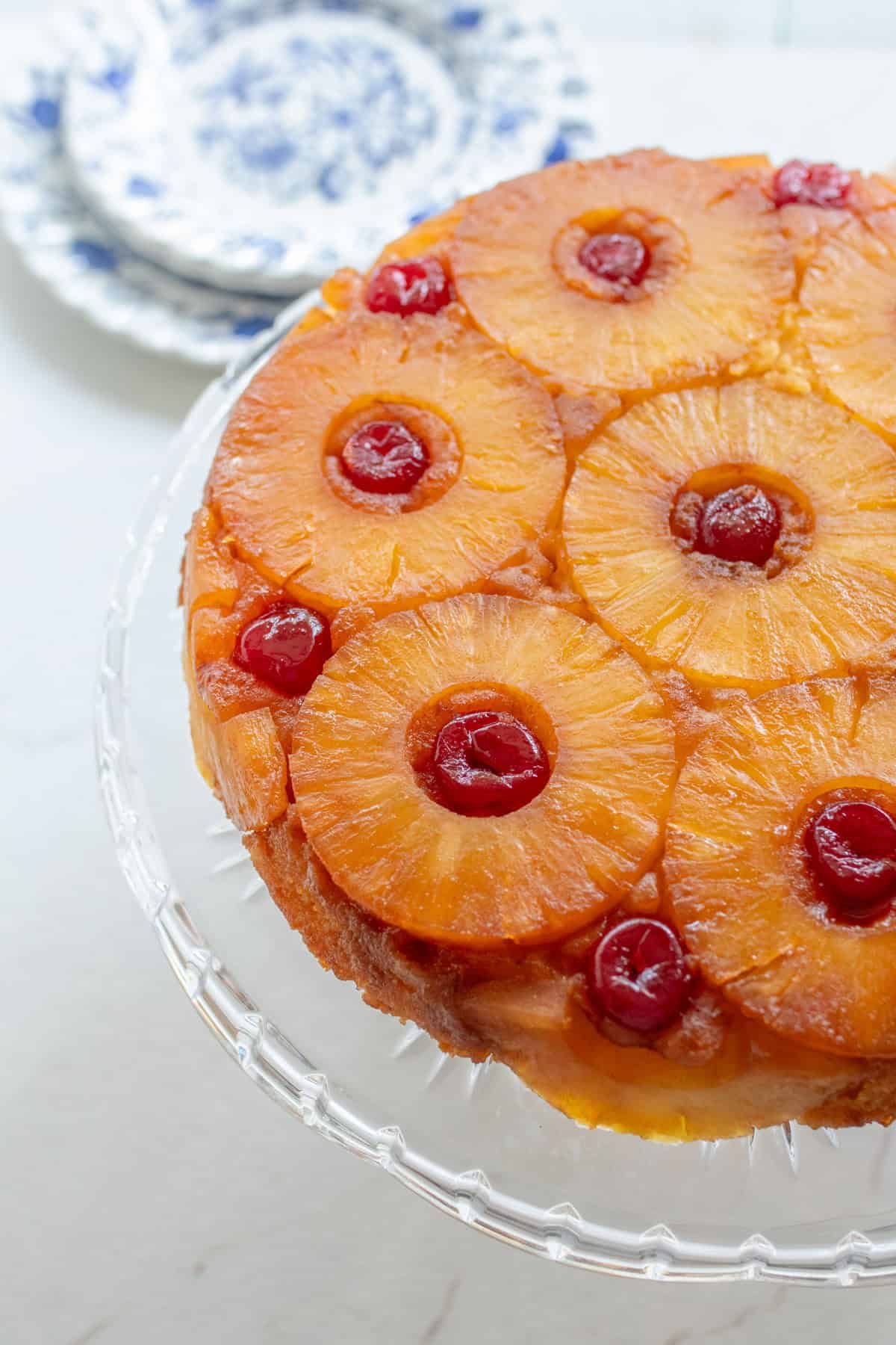 Close up picture of a pineapple upside down cake on a glass cake stand with plates in the background.