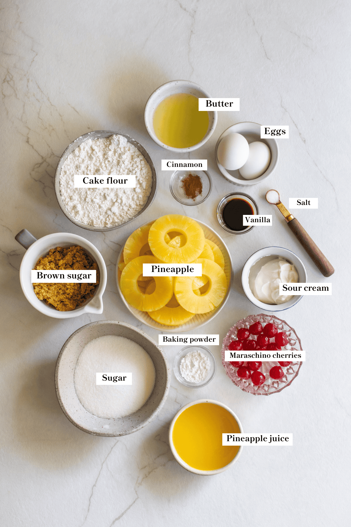 Pineapple, cherries, flour, eggs, sugar, and other ingredients in small bowls on a countertop.