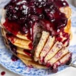 six fluffy pancakes layered on a plate drizzled with blueberry syrup.