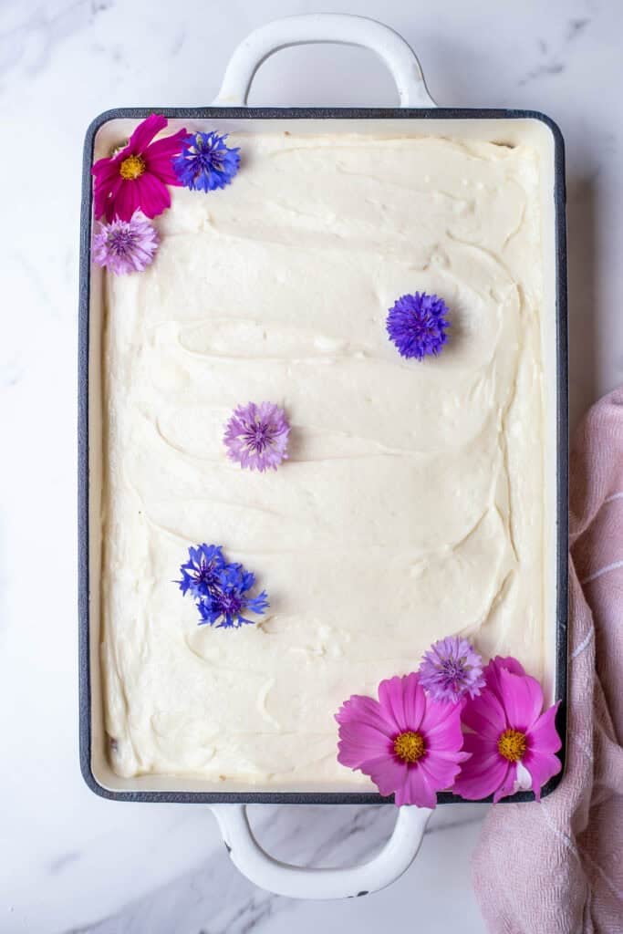A cake iced with white cream cheese frosting and decorated with pink and purple flowers.