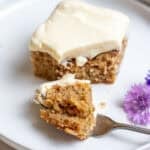 A bite sized piece of cake sitting on a white plate with a fork underneath.