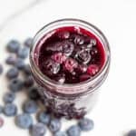 a glass jar full of blueberry sauce with several blueberries around the jar on the counter.