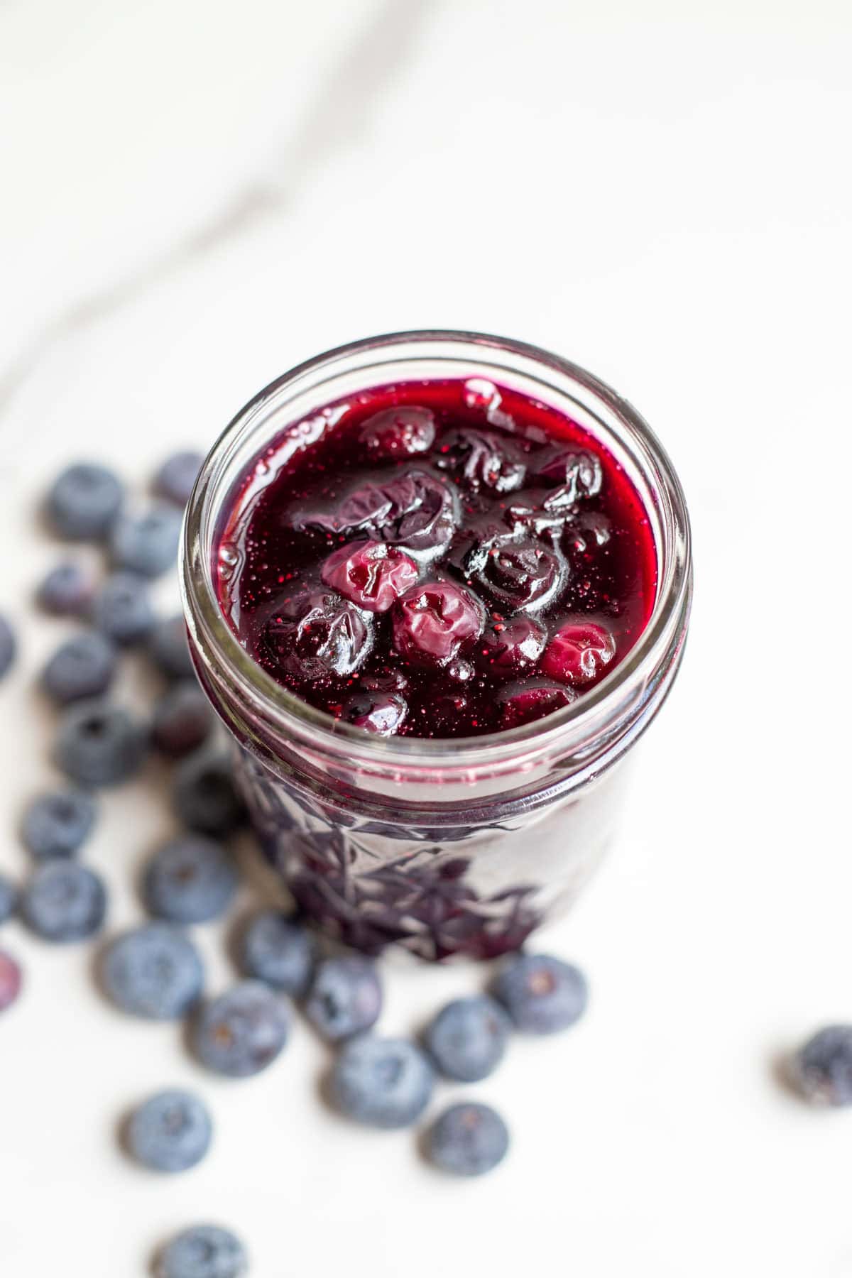 A glass jar full of blueberry sauce with several blueberries around the jar on the counter.