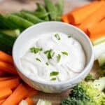 a white bowl of ranch dip surrounded by vegetables.
