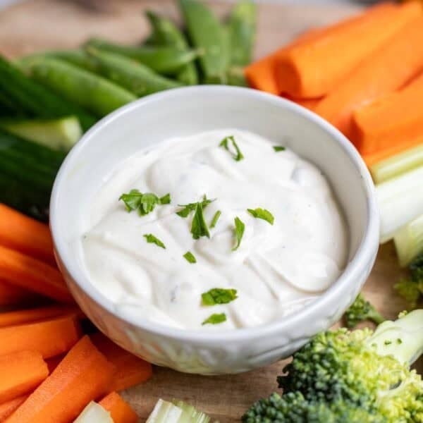 a white bowl of ranch dip surrounded by vegetables.