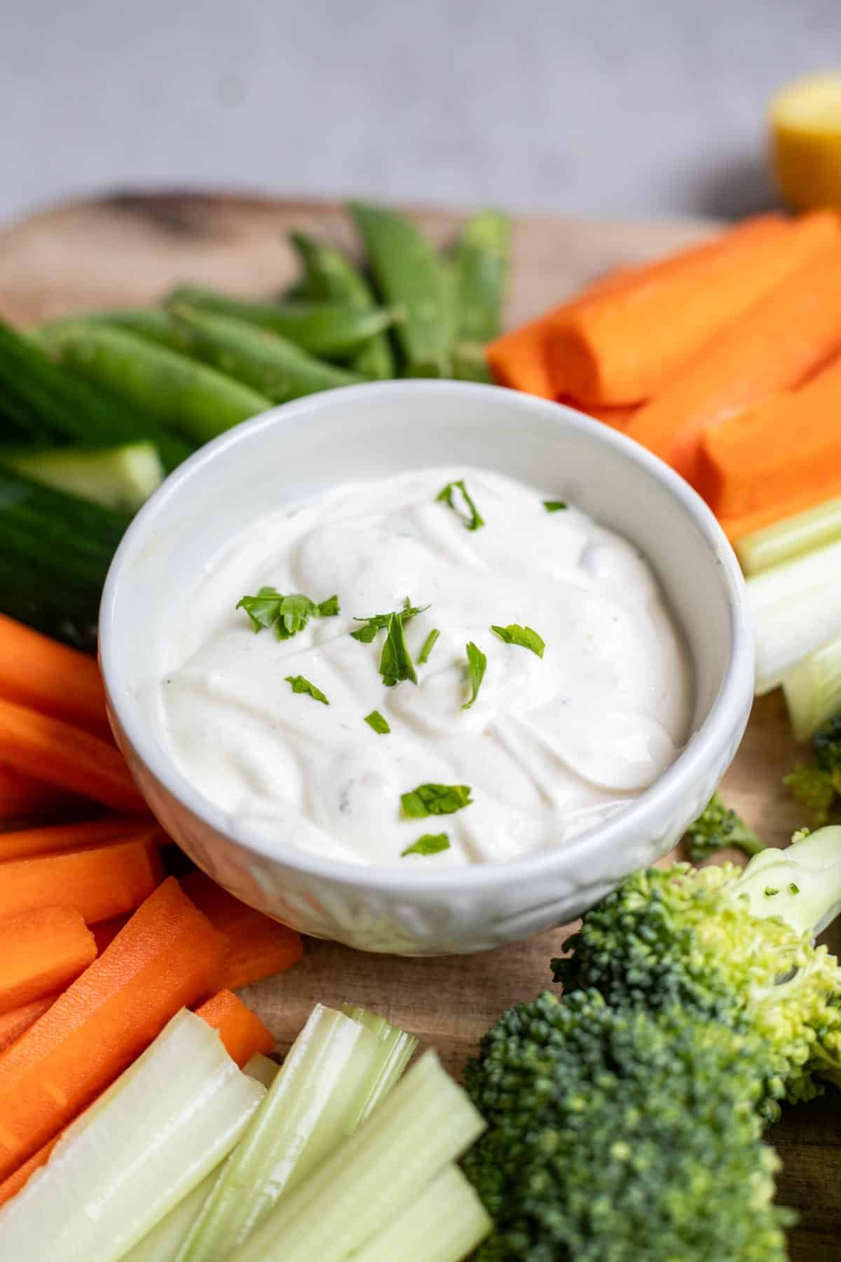 A white bowl of ranch dip surrounded by vegetables.