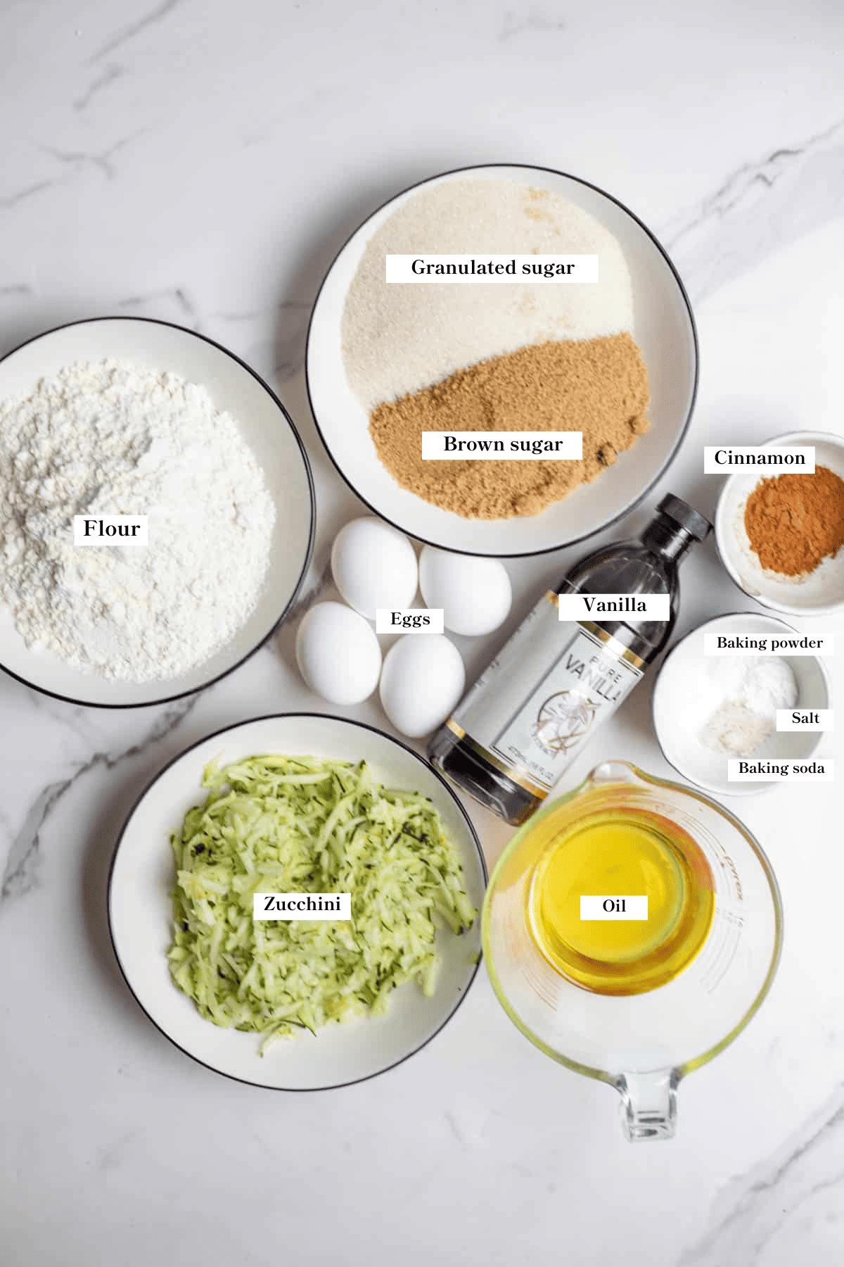 Zucchini, flour, sugar, eggs, oil, vanilla, and spices on the countertop.