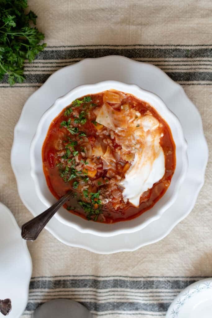 Cabbage soup served with sour cream and fresh herbs in a white bowl on a white plate.