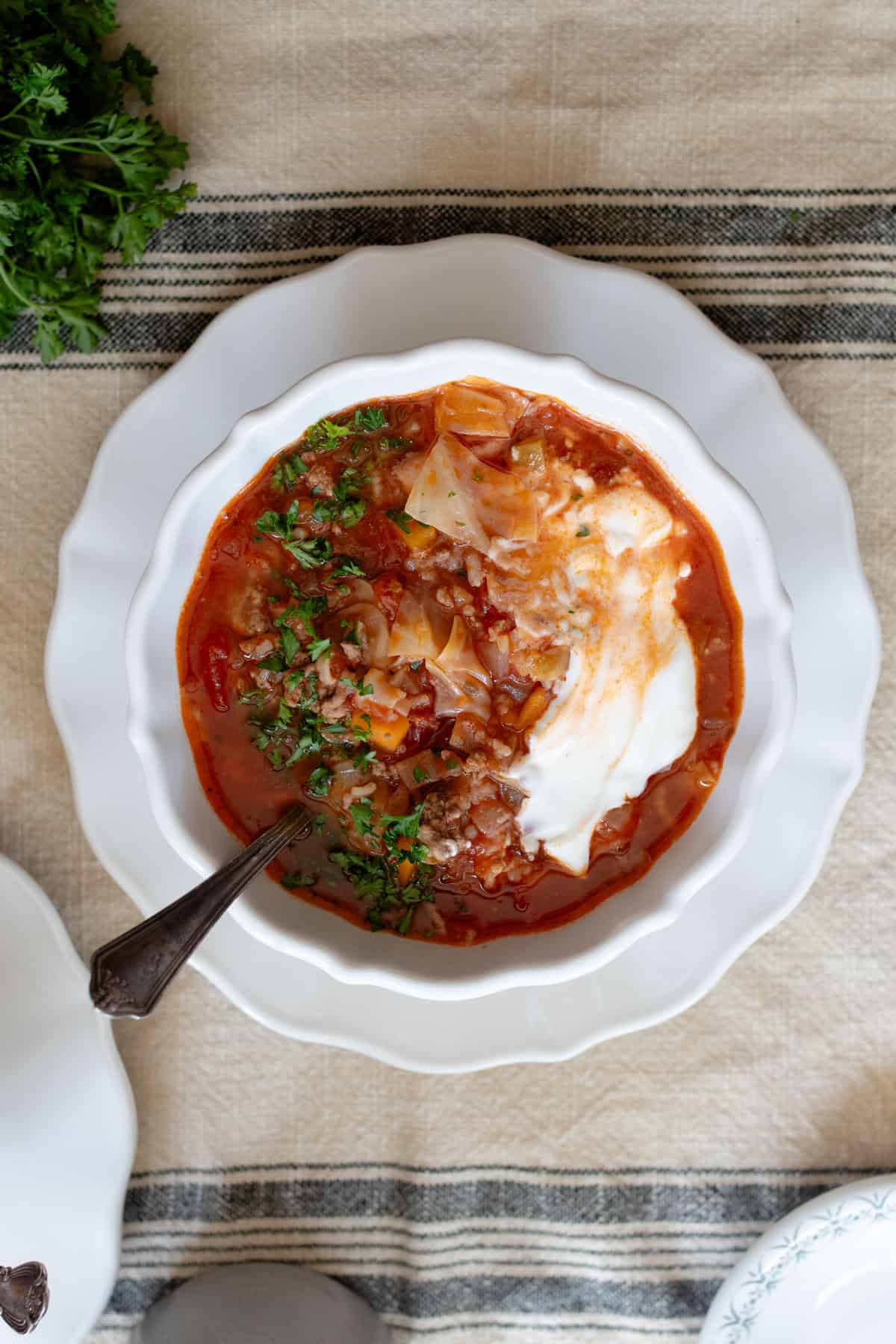 Cabbage soup served with sour cream and fresh herbs in a white bowl on a white plate.