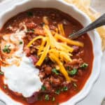 Ground beef chili topped with cheese, sour cream and fresh herbs in a white bowl on a whit plate.