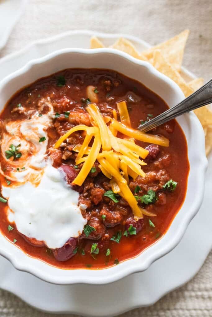 Ground beef chili topped with cheese, sour cream and fresh herbs in a white bowl on a whit plate.