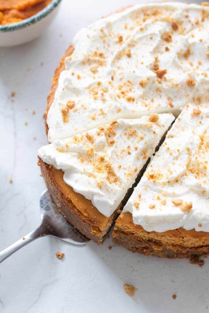 A pie server taking a slice of pumpkin cheesecake topped with whipped cream and gingersnap crumbs.