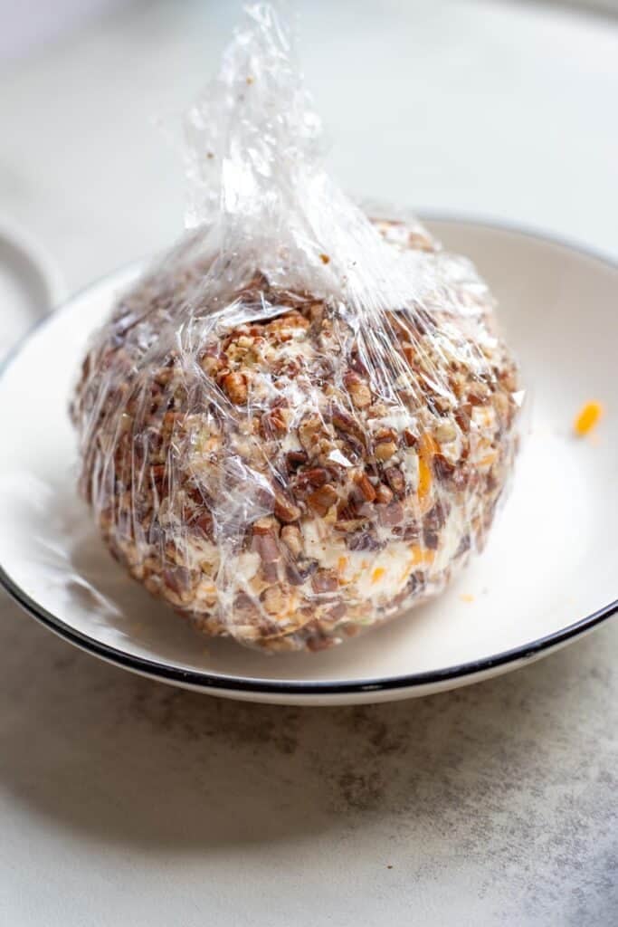 A cheese ball covered in pecan pieces and wrapped tightly in plastic wrap.