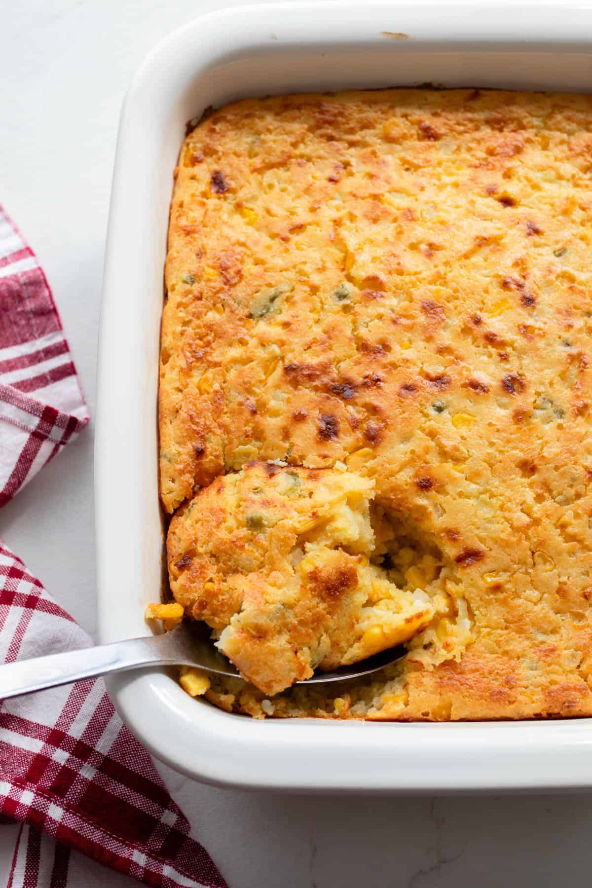 Half of a white baking dish full of creamy corn casserole with a spoon full of the casserole being lifted out.