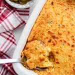 Corn casserole with a spoonful of casserole being scooped out of the baking dish. A red and white plaid towel and a small bowl of jalapeños sit next to the baking dish.