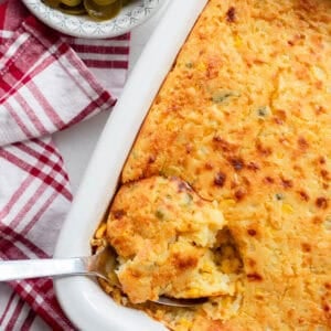 Corn casserole with a spoonful of casserole being scooped out of the baking dish. A red and white plaid towel and a small bowl of jalapeños sit next to the baking dish.