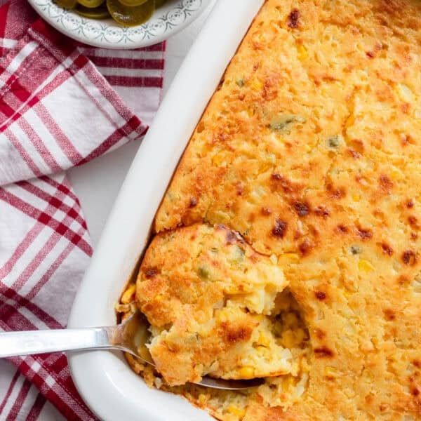 Corn casserole with a spoonful of casserole being scooped out of the baking dish. A red and white plaid towel and a small bowl of jalapeños sit next to the baking dish.