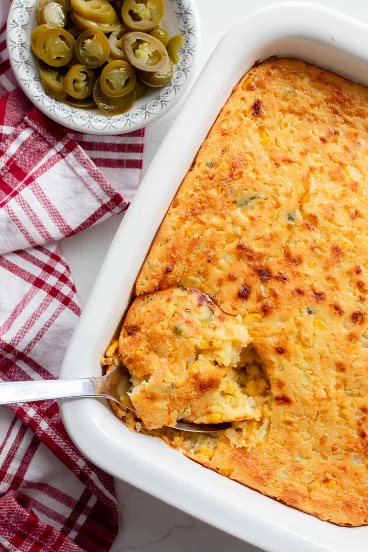 Corn casserole with a spoonful of casserole being scooped out of the baking dish. A red and white plaid towel and a small bowl of jalapeños sit next to the baking dish.