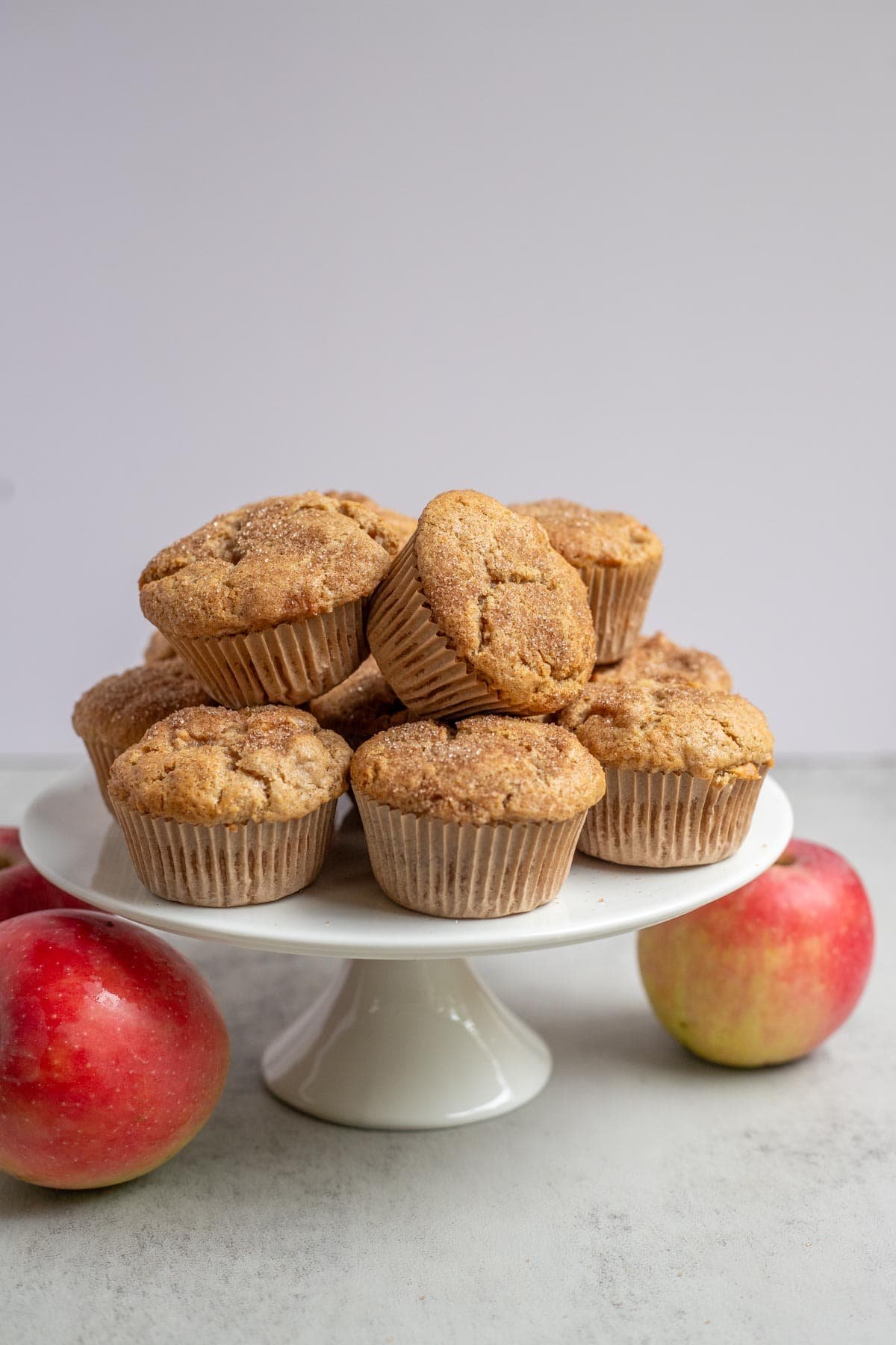 8 muffins stacked on a white pie plate with two red apples on the counter next beside it.