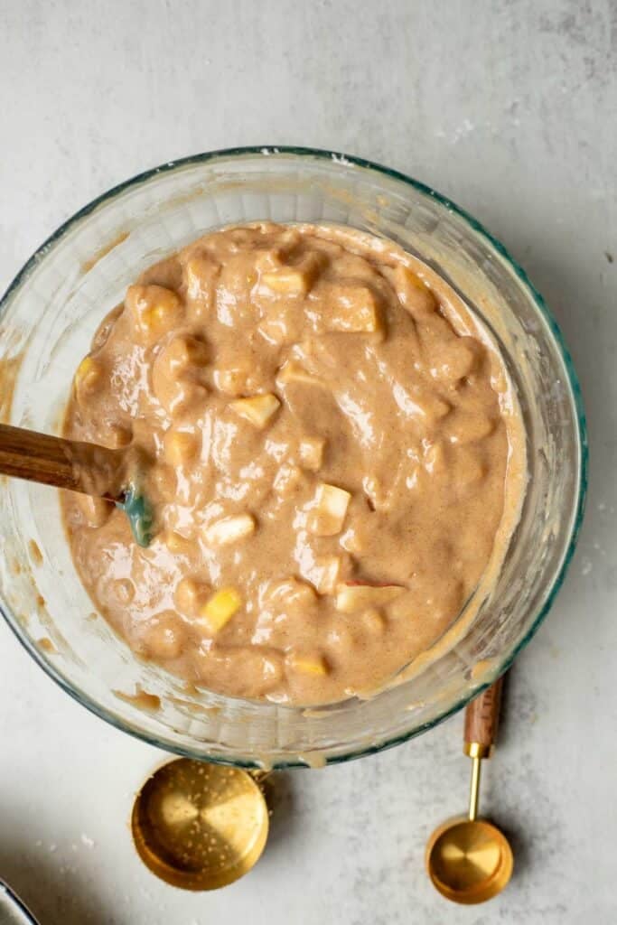 Apples in a muffin batter with a mixing spoon in the bowl.
