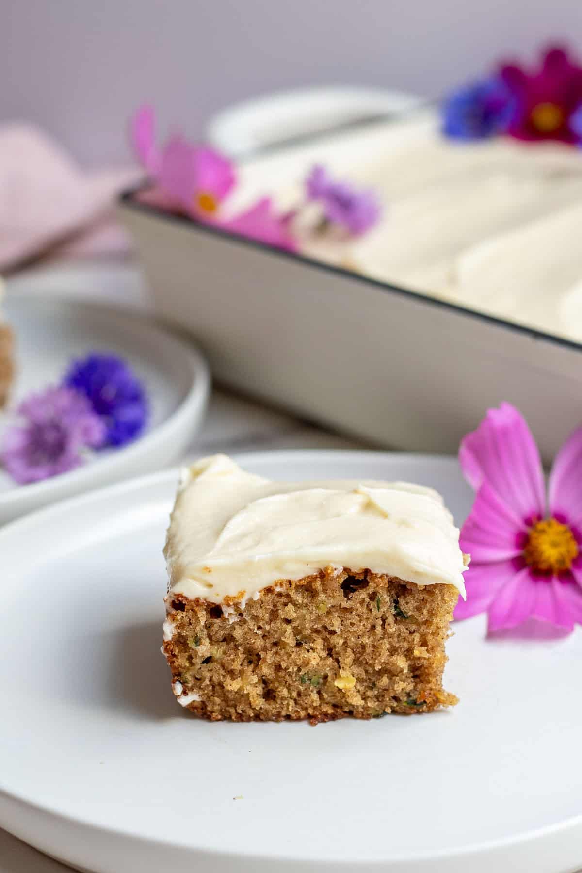 A slice of cake with cream cheese frosting sitting on a white plate with a pink flower beside it.