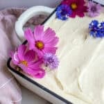A 9x13 cake covered with cream cheese frosting with flower petals used to decorate the cake.