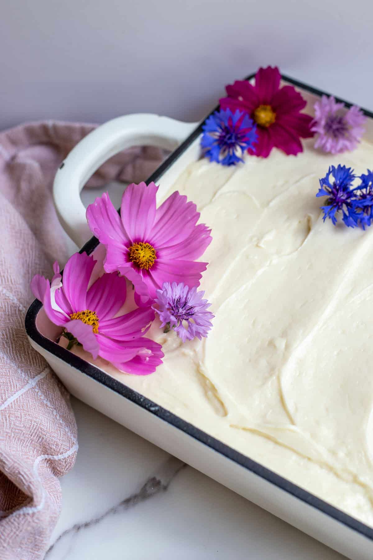 A 9x13 cake covered with cream cheese frosting with flower petals used to decorate the cake.
