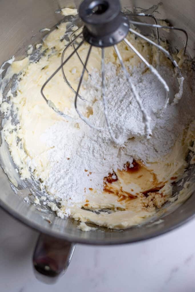 Powdered sugar and vanilla in a metal mixing bowl with whipped butter and cream cheese.